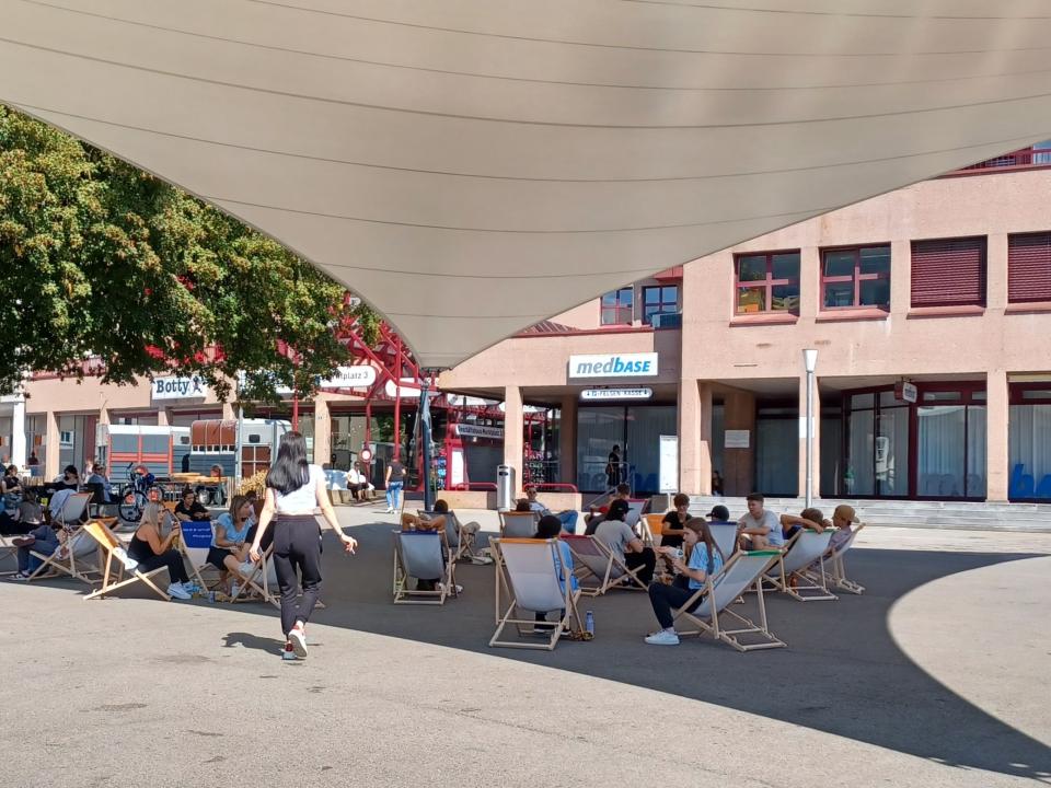 Unter einem grossen Sonnensegel auf einem belebten Platz sitzen zahlreiche Menschen entspannt auf bunten Liegestühlen. Im Hintergrund sind Geschäfte und ein Baum zu sehen. Die Szene wirkt einladend und ruhig – ein Moment der Pause mitten in der Stadt.