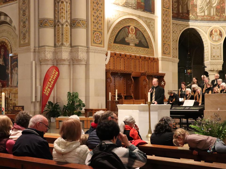 Chor singt in einer katholische Kirche, das Publikum sitzt auf Bänken und applaudiert.